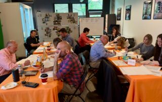 Group of professionals from Knauf, Fungi Media, Tell Your Story, and the Asthma & Allergy Friendly® Certification Program seated at orange-covered tables during a collaborative workshop.