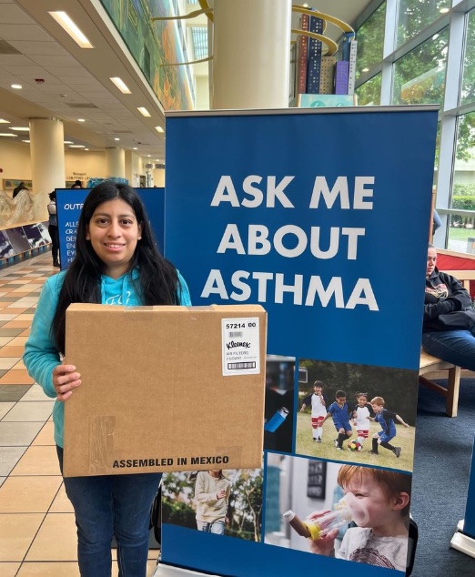 Community member holding a Kleenex® Certified air filter box in front of “Ask Me About Asthma” display.