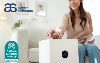 A woman adjusting a compact portable air purifier in her living room, representing the Asthma & Allergy Friendly® Certification Program’s updated standard for smaller certified air cleaners.